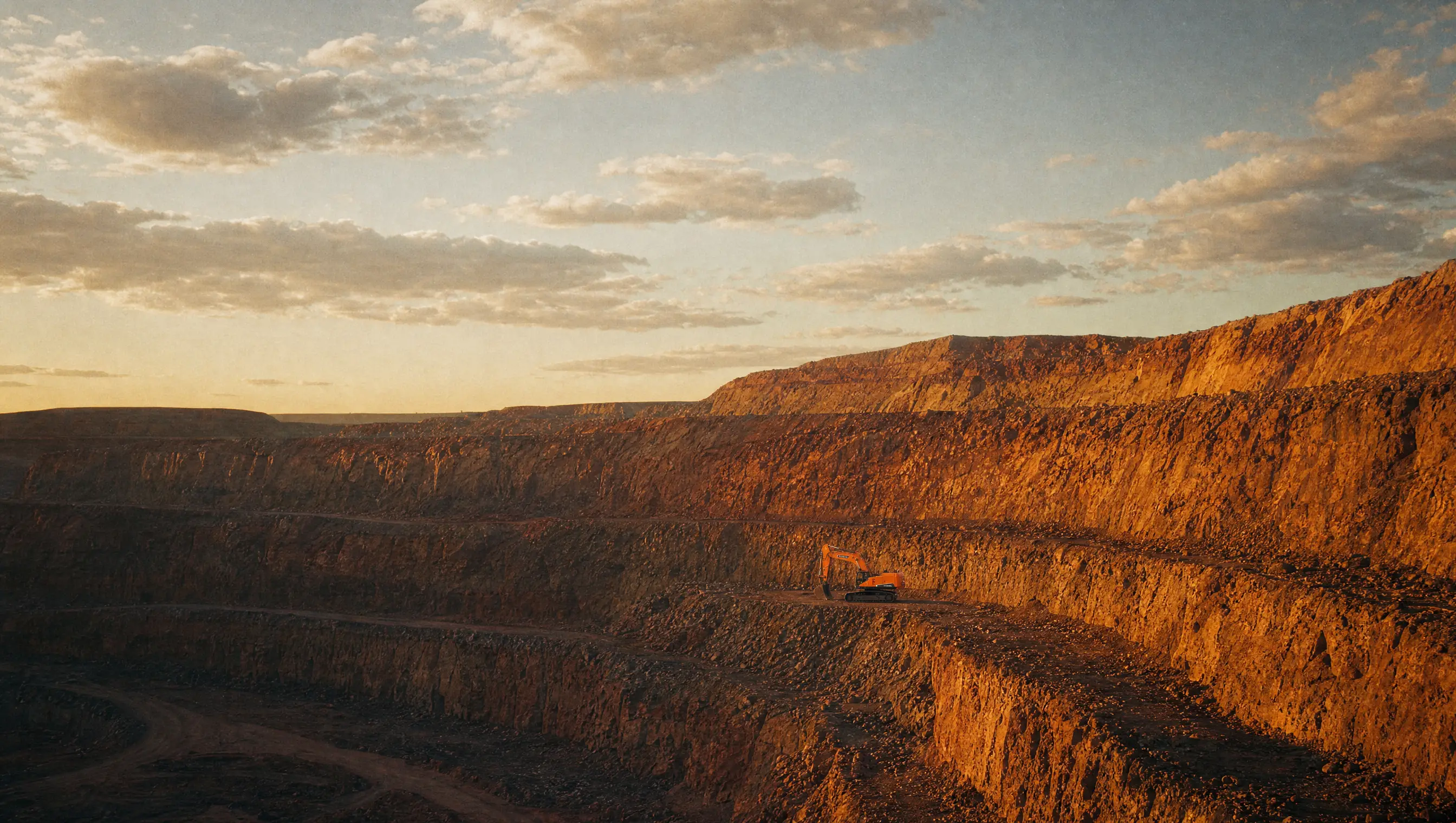 A wide benched open pit at golden hour, an excavator small in frame for scale under a vast African sky